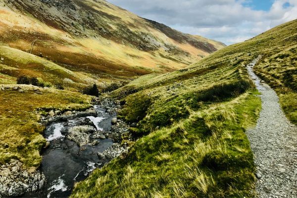 Narrow walking trail winding beside a mountain stream through a rugged moorland valley on the Coast to Coast walk, English Lake District